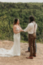 Bride in white gown holds hands with groom in suspenders on a rocky cliff, surrounded by lush green forest, creating a romantic scene.