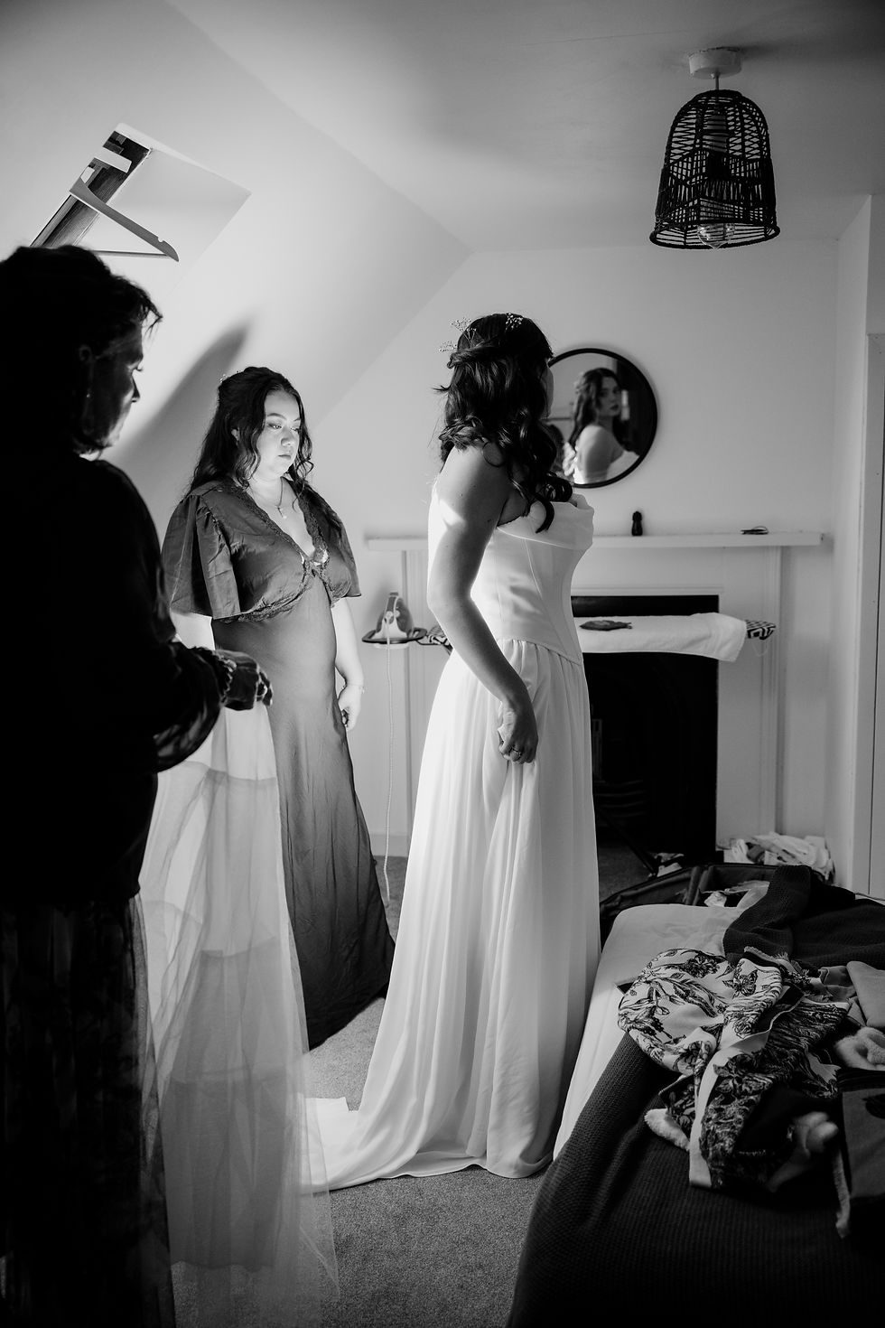 Black-and-white scene of three women in a bedroom, one in a wedding gown, others assisting, with a mirror and clothes on the bed.