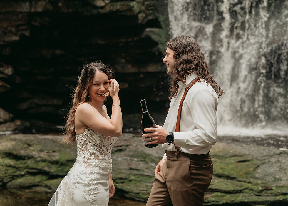 Couple smiles by a waterfall; woman in a lace dress, man in white shirt with suspenders holding a bottle. Joyful, natural setting.