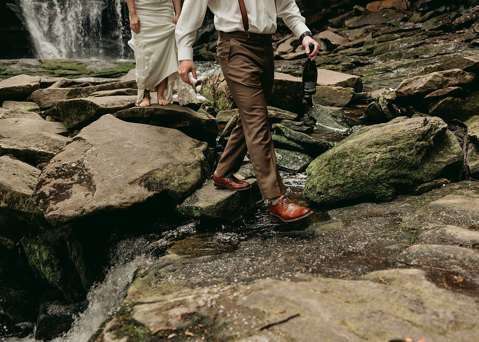 Couple in wedding attire crosses mossy rocks by a waterfall; groom holds a champagne bottle, creating a romantic, adventurous mood.