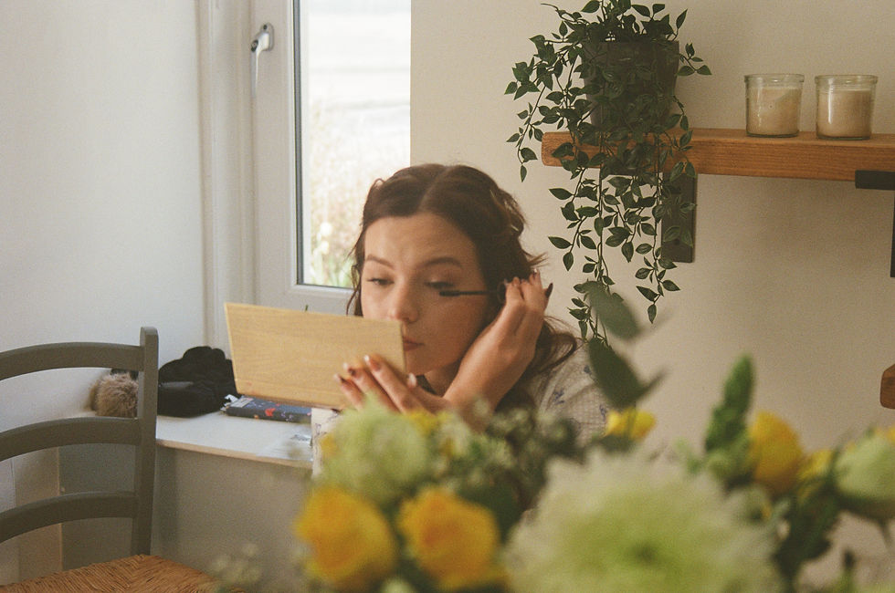 A film photo of a bride applying makeup with a hand mirror in a cozy room with plants, candles, and blurred yellow flowers in the foreground.