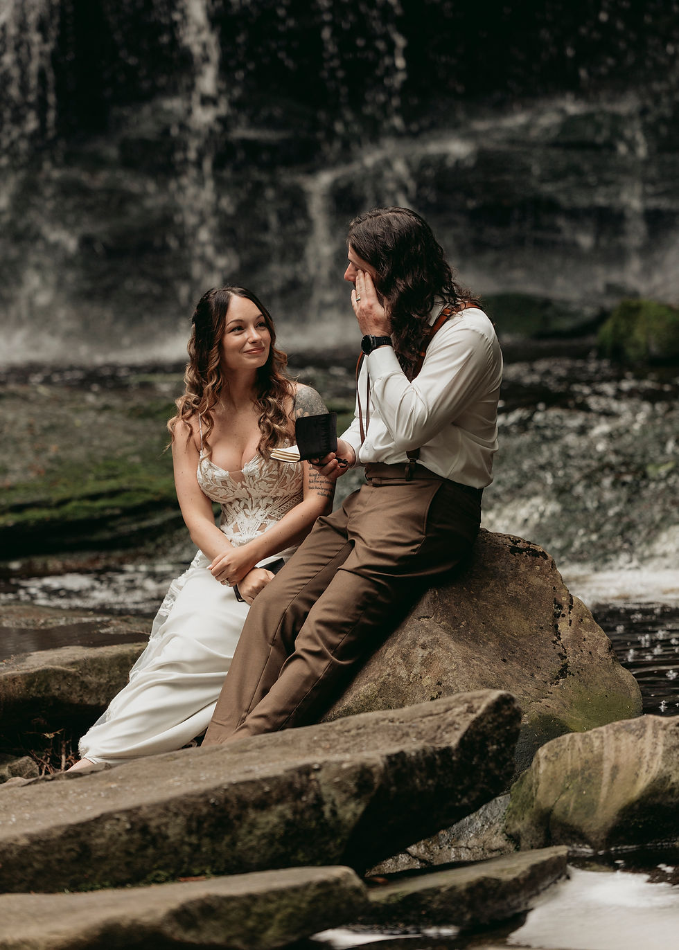 A couple sits on rocks by a waterfall. The woman smiles in a white dress, the man wipes a tear, holding a black book. Serene setting.