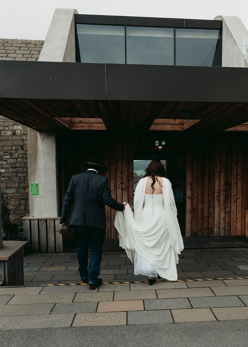 Bride in white dress and man in dark suit walk into a modern wooden building, seen from behind, calm wedding mood