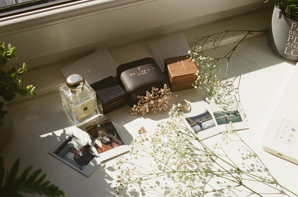 A film photo of a sunlit windowsill still life with perfume bottle, jewelry boxes, ring, Polaroids, and baby's breath flowers.