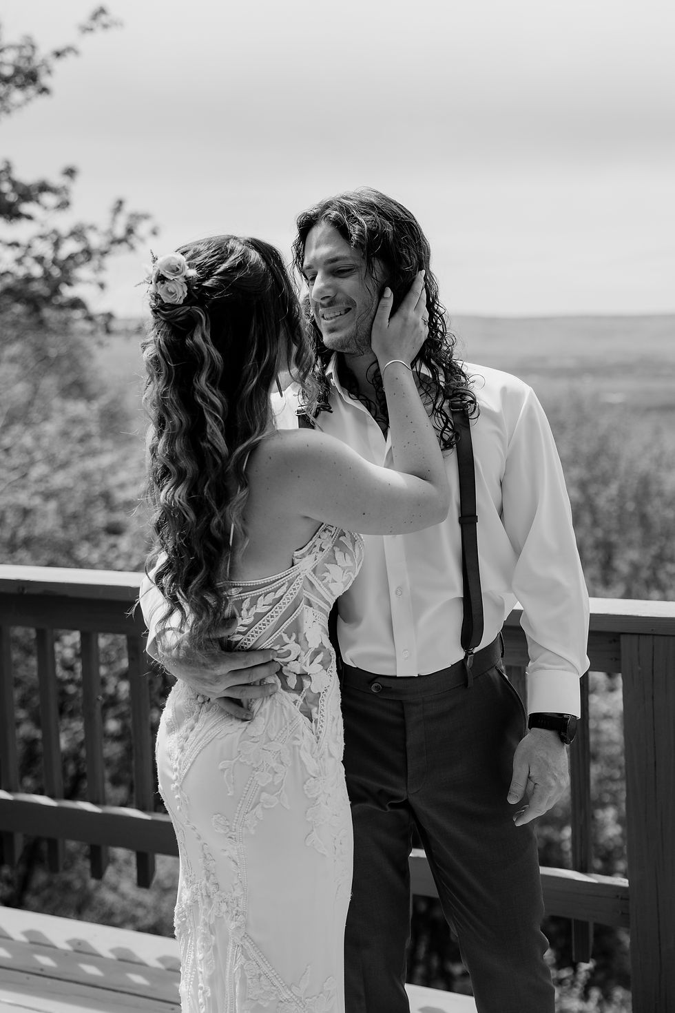 Bride and groom embrace on a balcony, overlooking a scenic landscape. Bride in lace dress, groom in shirt with suspenders. Romantic mood.