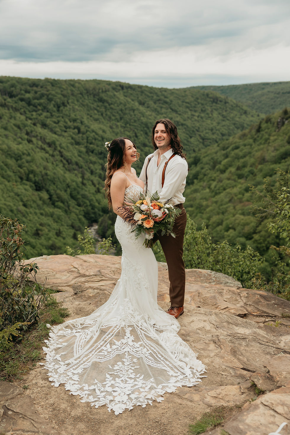 Bride in a lace gown holds a bouquet, laughing with the groom on a rocky overlook. Green forested valley under a cloudy sky.