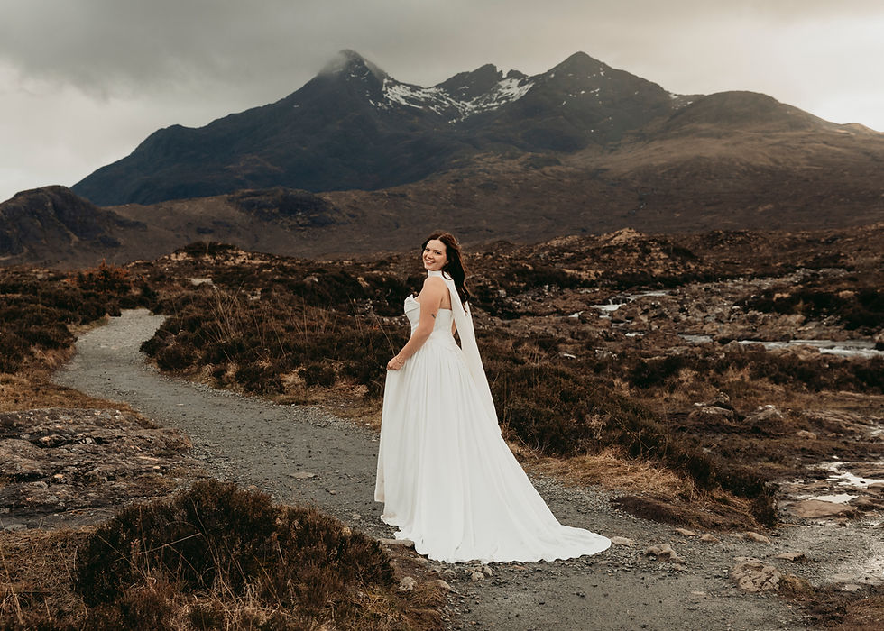 Smiling bride in a white dress stands on a rocky mountain path under cloudy peaks, with a moody landscape behind her.