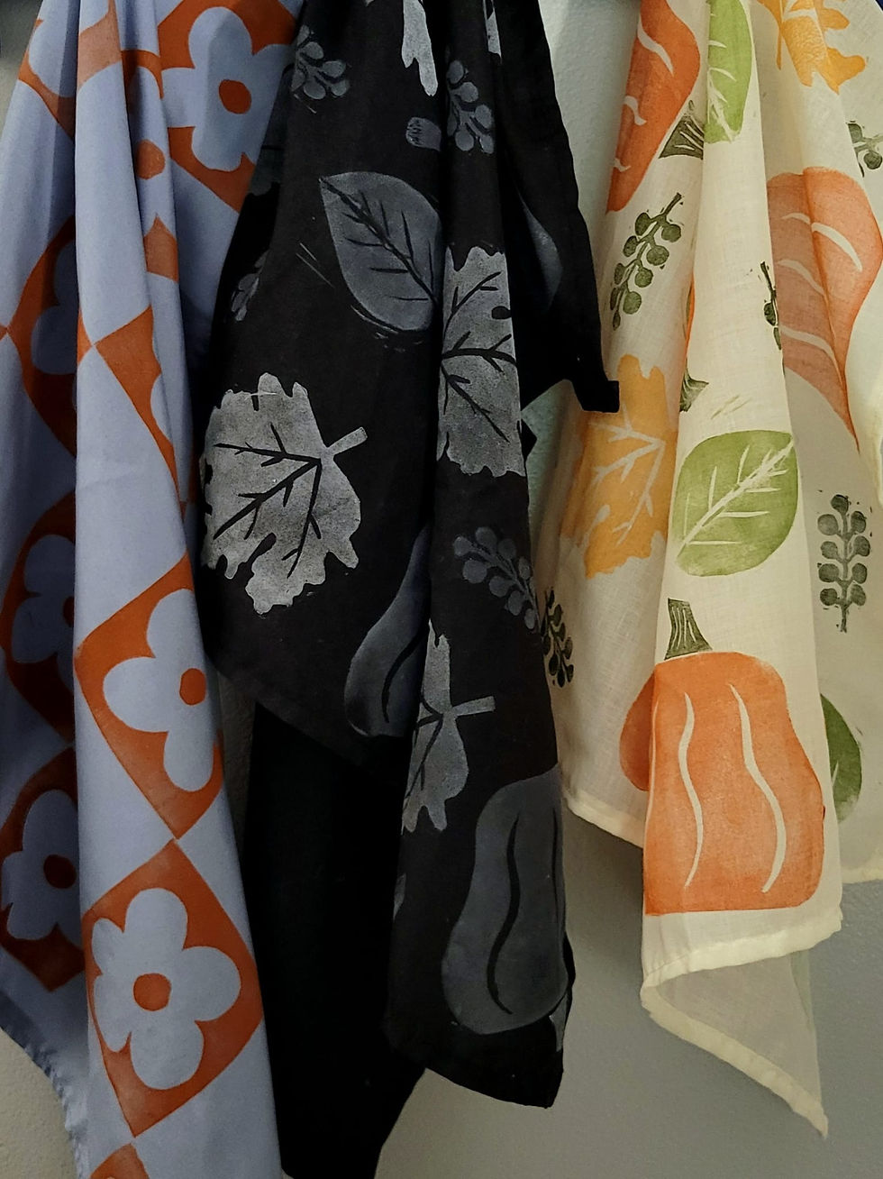 Three patterned bandanas hanging: one blue with orange flowers, one black with white leaves, and one cream with colorful leaves and pumpkins.
