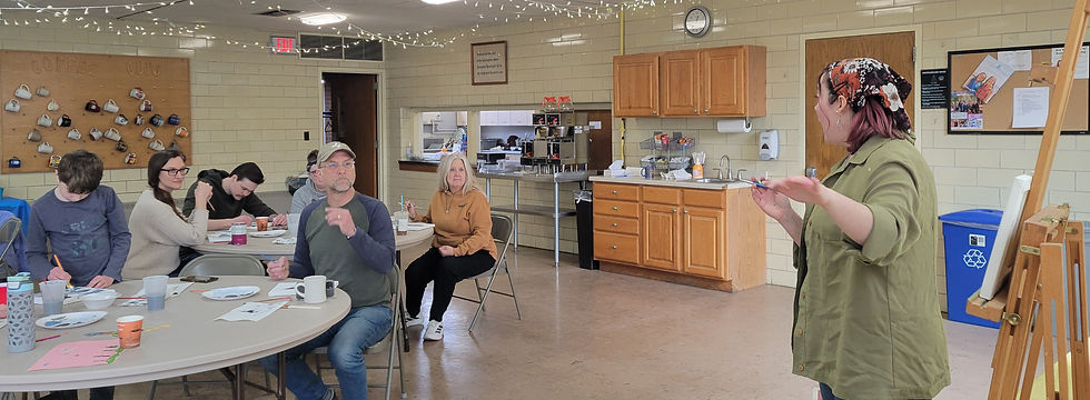 A group listens to a speaker in a cozy room with art supplies. Mugs adorn the wall, and a kitchen area is in the background