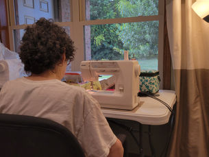 My Grandma sewing on a machine in a cozy room with a large window, facing a green garden. A fan, lamp, and teal-patterned bag are nearby.