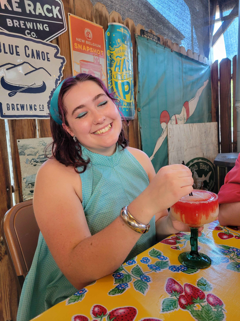 Artist Gwena Dye Pringle celebrates her 23rd birthday while holding a frozen strawberry margarita in a green-stemmed glass. She's wearing a handmade 60's inspired turquoise halter top with a textured pattern and has purple highlights in her dark hair. Gwena is smiling warmly while seated at a yellow tablecloth decorated with strawberry and blueberry prints. The restaurant setting features vintage brewery signage including Blue Canoe Brewing Co. and New Belgium Snapshot signs, along with colorful beer cans and eclectic decor in the background, creating a fun, retro atmosphere for her birthday celebration.