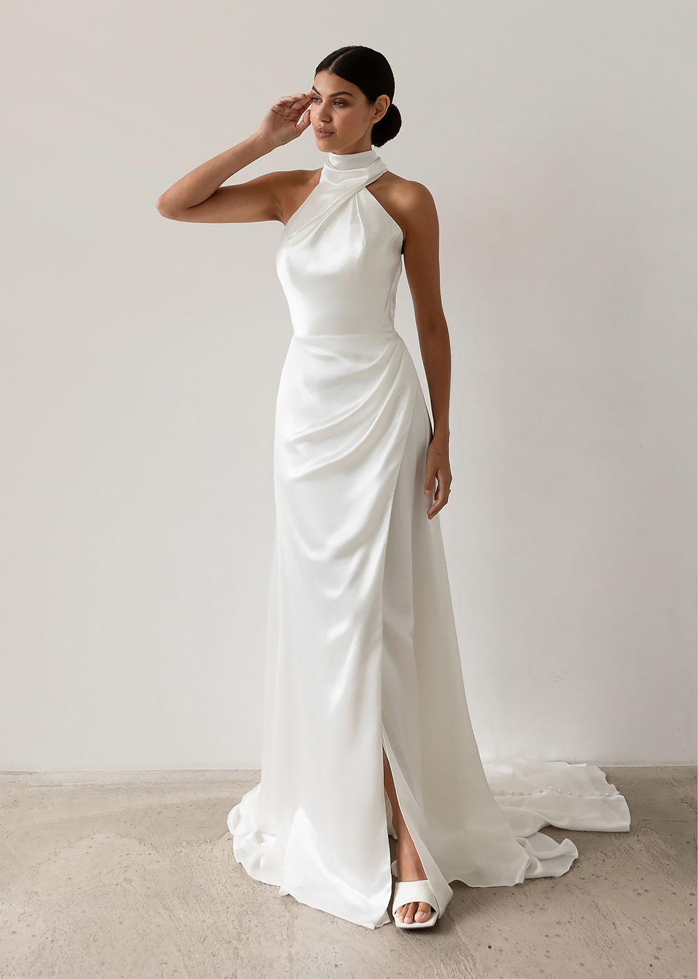 A woman in a sleek, white halter-neck satin wedding gown poses confidently against a plain backdrop, showcasing elegance and sophistication. Available at The Corner Atelier, Manchester.