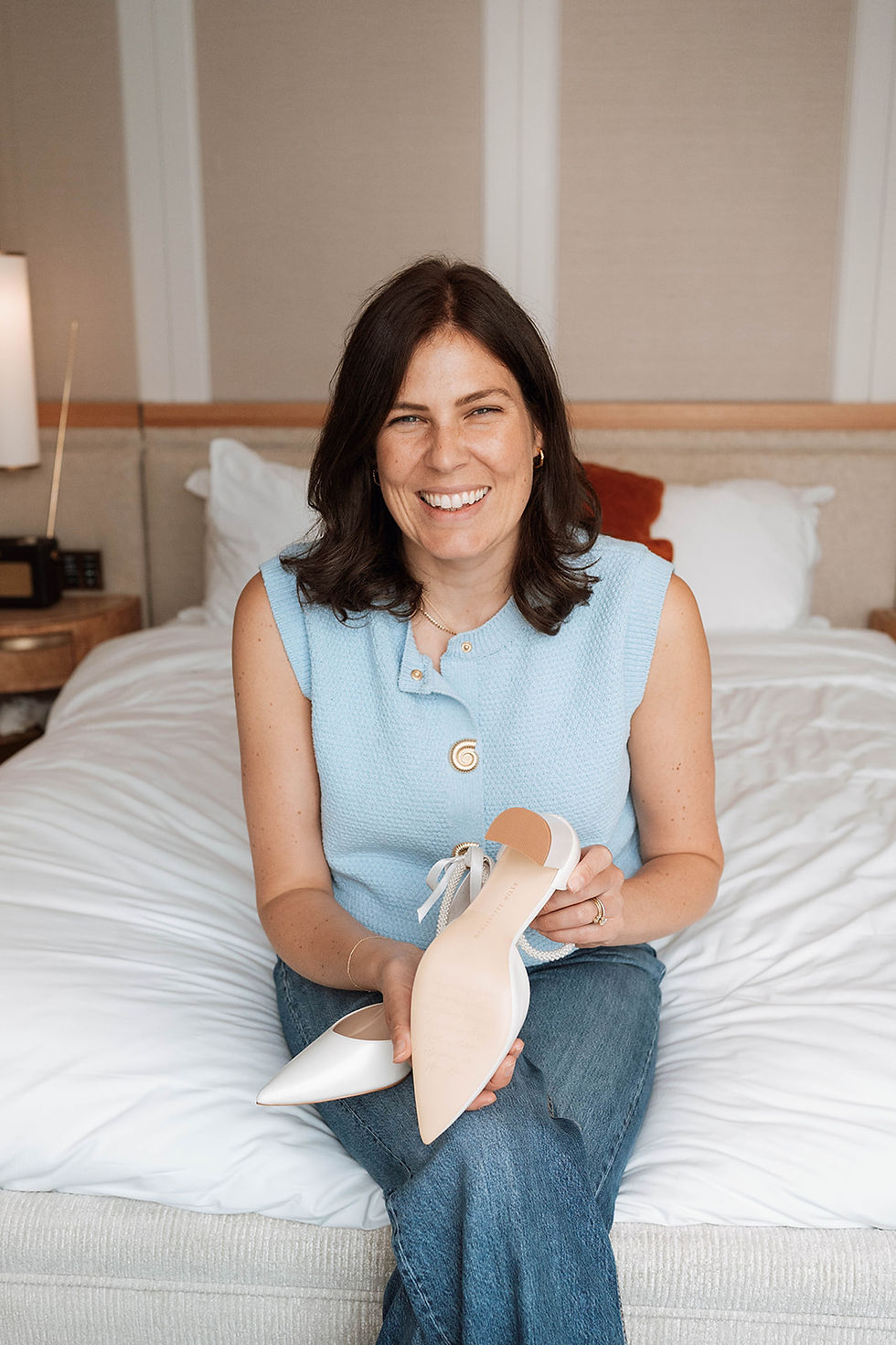 Smiling Charlotte Mills in light blue top and jeans, sitting on a bed, showcasing her beautifully designed bridal shoes in a cozy room with white bedding.