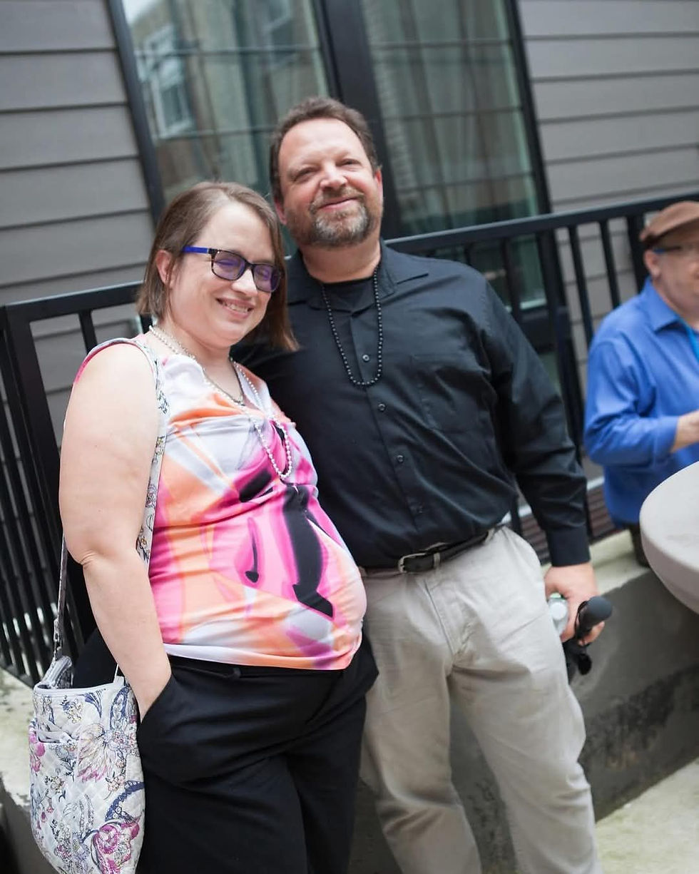 Smiling man in black shirt and woman in colorful top stand together outdoors. Another person in blue shirt and hat is blurred in the background.