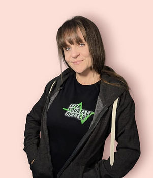 A photo of Tonia standing with her body slight rotated away from the camera, looking at the camera. She has brown hair which is long with some golden tones. She is wearing a black t-shirt whcih has the Live Yourself Better lofo on the right side as we view it. She is also wearing a grey hoodie
