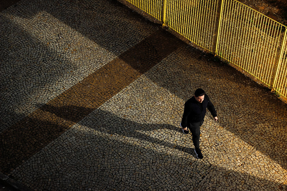 Homem de preto caminha em calçada de pedras sob sombra longa. Cerca amarela ao fundo. Atmosfera de fim de tarde com luz suave.