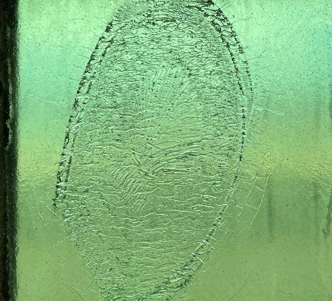 Visitors to the Museum of American Glass will encounter a site-specific window intervention by guest artist and curator, Jocelyne Prince. Her work, Ghost Optics, addresses early methods of flat glass and bottle glass production. Of particular interest to the artist was the variety of Americana imagery found on the historic glass bottles in the collection. Wanting to source these images as directly as possible, Prince dug into the WheatonArts mold collection and honed in on one particular mold, a flask for spirits, which had an inset for interchangeable emblems. The imagery for these emblems was a mix of patriotic, historic, and commemorative motifs. This mold was part of an industrial technology known as the “American System.” Similar to industrial advances in other kinds of production, the glass industry capitalized on a bottle mold with interchangeable parts – in this case an assortment of emblem inserts meant that one mold could be used to make a multitude of different bottles.
Working with the Paul Wissmach Glass Company in West Virginia, Prince hot cast the various emblems and then fed them into the industrial glass roller. The emblems are stretched, crackled, and sometimes even fractured—their ghost-like watermark characteristic becomes visible at certain angles and invisible at other angles. This material process mirrors how we test our patriotism and democracy
. . . challenging its limits, and sometimes going beyond.