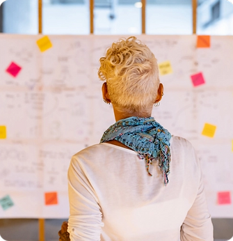 Person with short blonde hair and a scarf looking at a whiteboard filled with colorful sticky notes and sketches in a collaborative workspace.