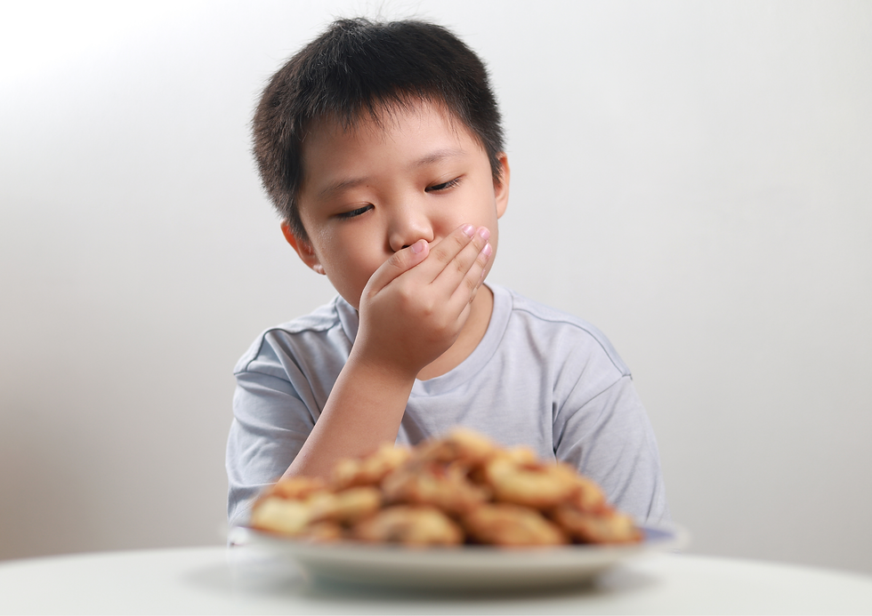Young boy covers his mouth and looks uneasily at a plate of food on a white table against a plain white background