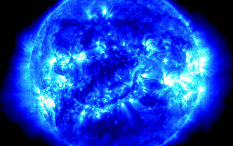 Image du Soleil en teintes bleues montrant sa couronne et plusieurs régions actives brillantes. Des structures lumineuses et tourbillonnantes de plasma sont visibles à la surface et autour du Soleil.

