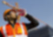 Construction worker standing outside wearing orange hardhat, goggles and orange reflective vest drink a plastic bottled water with his left hand.