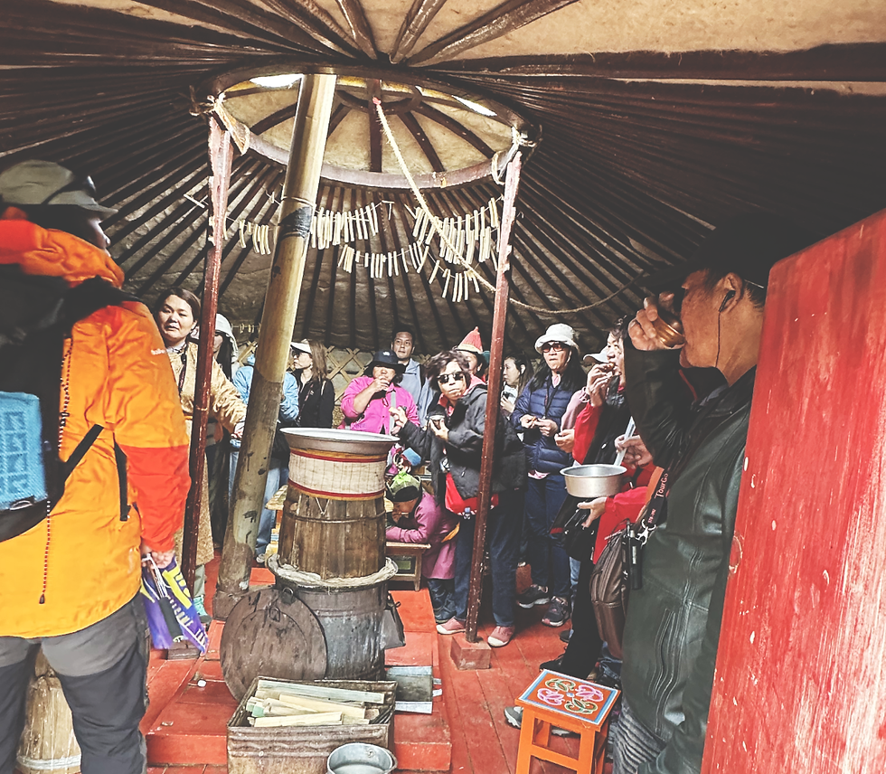 People gather inside a yurt, some wearing bright jackets. A person drinks from a bowl. Red and wooden interior with a festive, cozy atmosphere.