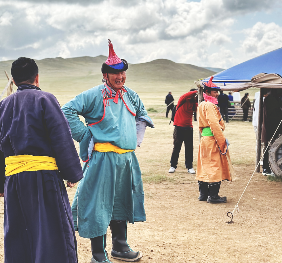 People in colorful traditional attire and hats converse outdoors, with tents and hilly grasslands in the background under a cloudy sky.