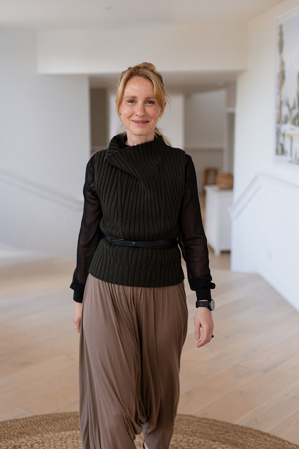Smiling woman in a black top and brown skirt stands in a bright minimalist room with white walls and a wood floor.