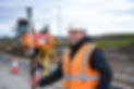 Young man smiling with hard hat and orange high vis jacket. He is on a construction site, with tools and there is a track machine in the background