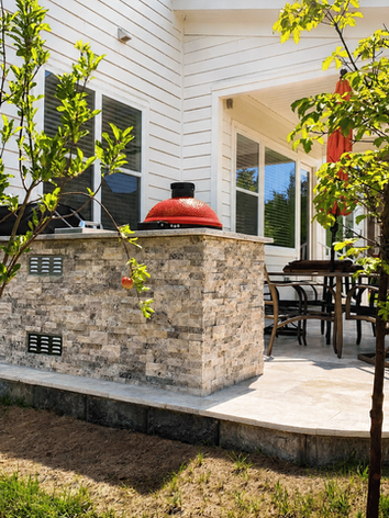 Stone outdoor kitchen with red ceramic grill, patio dining area, and backyard landscaping in Charlotte NC