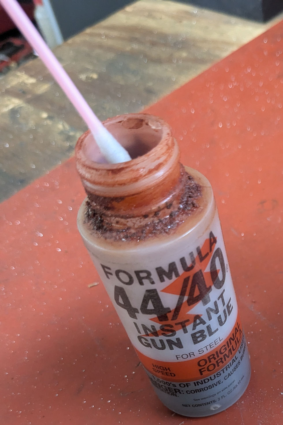 Open bottle of Formula 44/40 Instant Gun Blue on a workbench. A swab is inside the bottle. Text and orange label are visible.