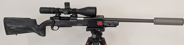 A sniper rifle with a silencer and scope mounted on a tripod against a plain background. Camouflage pattern with notable red detail.
