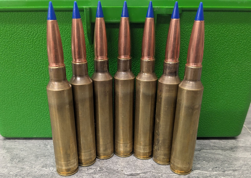 Seven brass bullets with blue tips stand upright in front of a green textured box on a gray surface. No text visible.