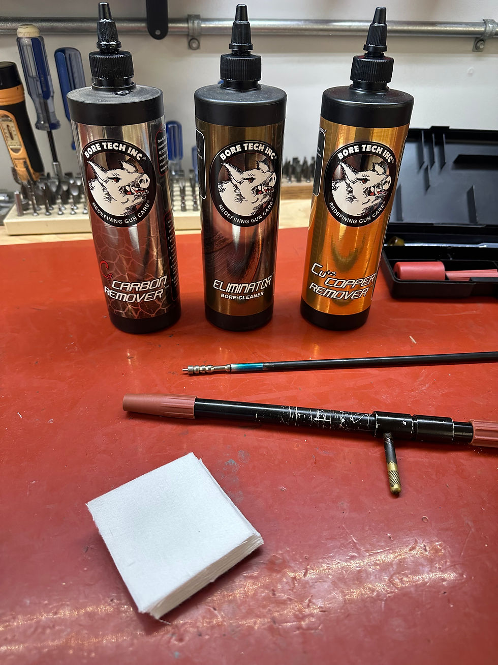 Three cleaning product bottles labeled "Bore Tech Inc" on a red table, with cleaning rods, white cloth, and a toolbox in the background.