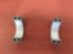 Two metal scope ring clamps with holes on an orange workbench. The scope ring caps are gray and curved, positioned with space between them.