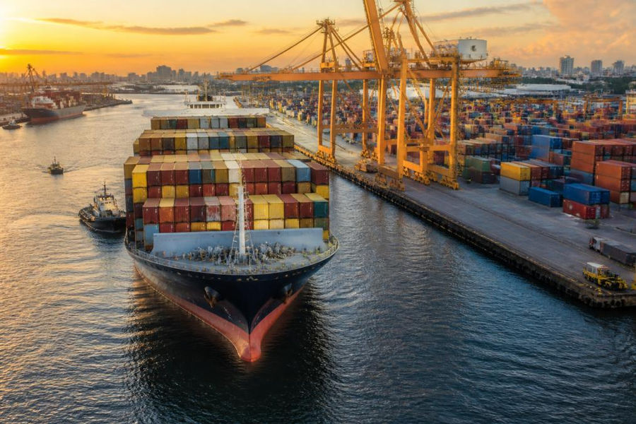 Aerial view of a large container ship loaded with stacked shipping containers entering a busy commercial port at golden hour, with cranes and dock workers visible