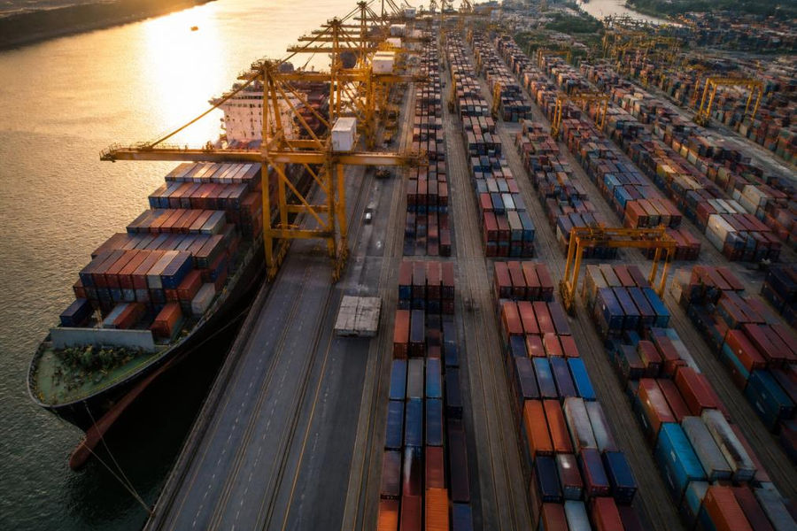 Aerial view of a busy container port at golden hour, rows of stacked 20 ft shipping containers in red and blue stretching toward a massive cargo ship being loaded by gantry cranes