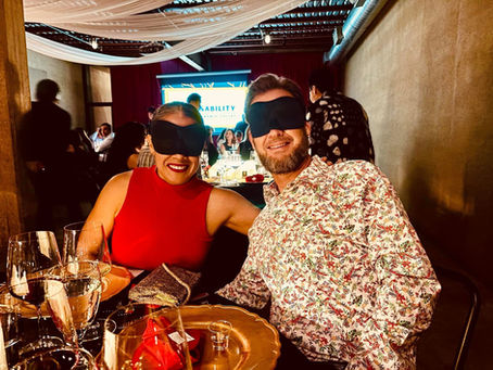A couple smiling at the camera while blindfolded at dinner. The woman is wearing a red dress, and the man is dressed in a multi-colored shirt.