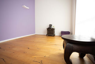 Auf dem Bild sieht man einen kleinen Yoga-Raum mit Holzboden. Die Font- und Seitenwände sind weiß, die linke Seitenwand ist lavendel. Das Bild ist von schräg rechts aufgenommen. Vor der Frontwand steht ein kleiner Holzaltar mit einem Ganesha. Rechts daneben liegen vier zusammengefaltete Decken. Am rechten Rand steht ein kleiner Holztisch.