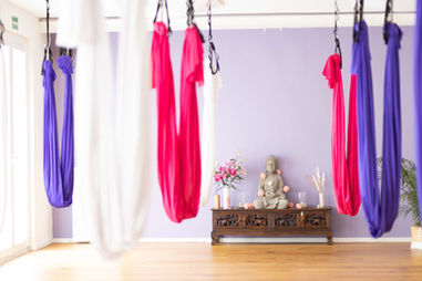 In einem hellen Raum mit lavendelfarbener Frontwand und Holzboden steht ein länglicher, niedriger Holzaltar vor der Wand. Von der Decke hängen verschiedenfarbige Aerial-Yoga Tücher.