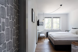 Das Bild zeigt ein Doppelzimmer. Die Fenster vor Kopf und die weißen Wände machen den Raum hell und freundlich. Rechts im Bild steht ein Doppelbett. Der Boden ist aus Holz. Im linken Bildvordergrund ist ein Stück einer schwarz-weißen Tapete, die die Diele auskleidet erkennbar.