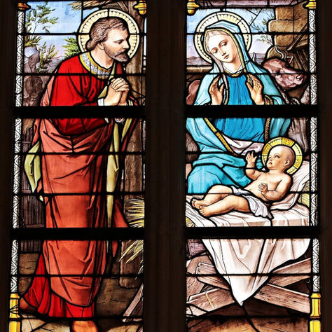 Detailaufnahme eines Kirchenfensters mit Weihnachtsmotiv, farbige Glasmalerei mit Heiligenfiguren und Kind in der Krippe.