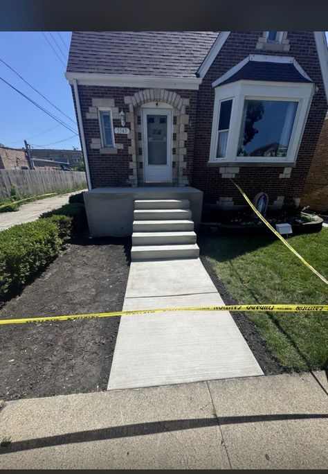 Front view of a newly installed concrete sidewalk and front porch steps with a fresh finish, set against a classic red brick house with stone accents, cordoned off by yellow caution tape.