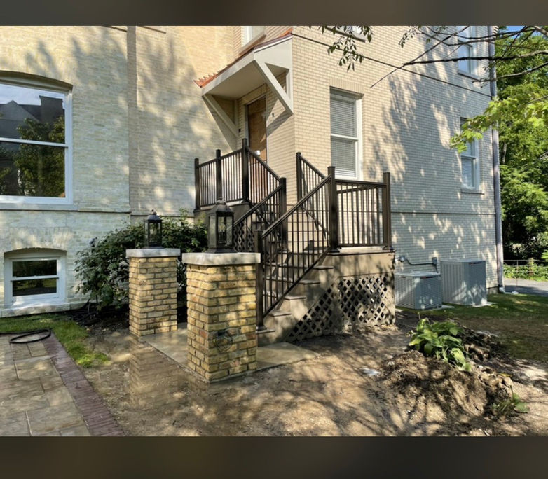 A finished residential entryway featuring two custom yellow brick pillars with limestone caps and decorative lanterns, leading to a porch with black metal railings and a decorative lattice base