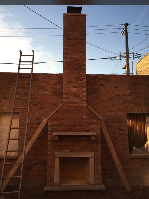 An outdoor construction site showing a custom-built red brick fireplace and full chimney under a clear sky, featuring a finished firebox, a heavy stone mantel, and structural timber bracing for temporary support