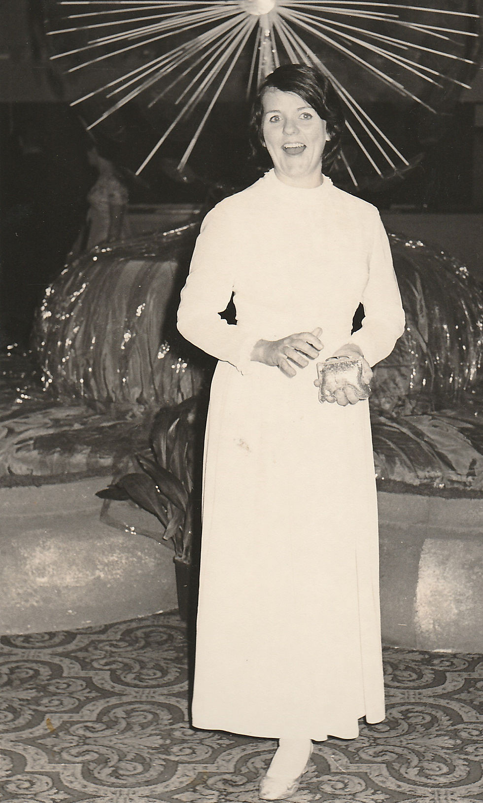 A black and white photograph of a young woman, about 25 years old, dressed in a long-sleeve white gown, standing indoors with an amused expression. She is clapping her hands lightly and looking slightly to her left. Behind her is a decorative, fan-like backdrop, enhancing the festive ambience of the scene.