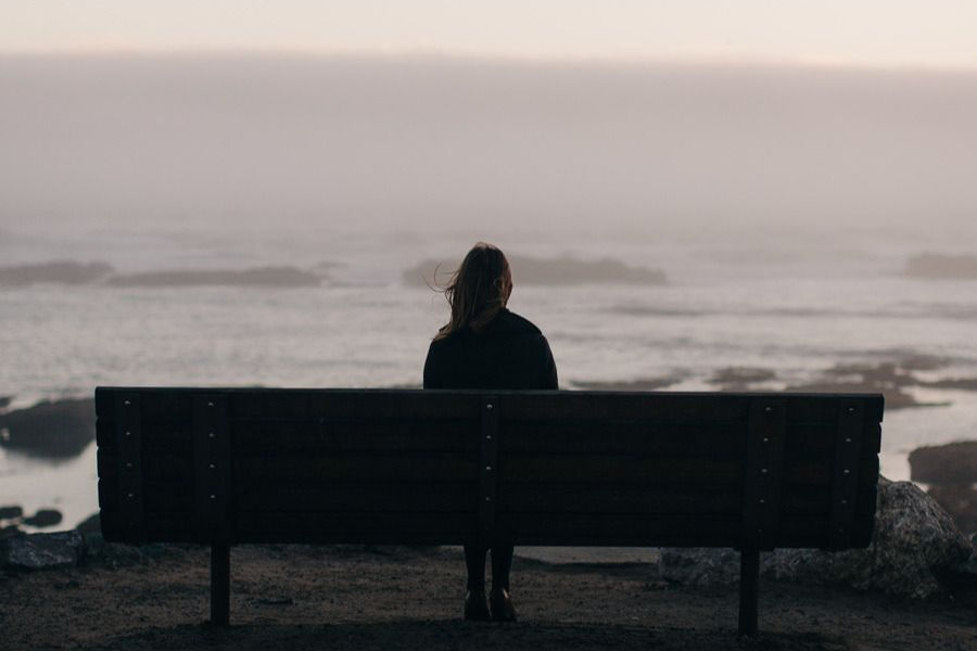 A woman sits alone on a bench by the sea, quietly reflecting — a moment of solitude after surviving what no one else could see.