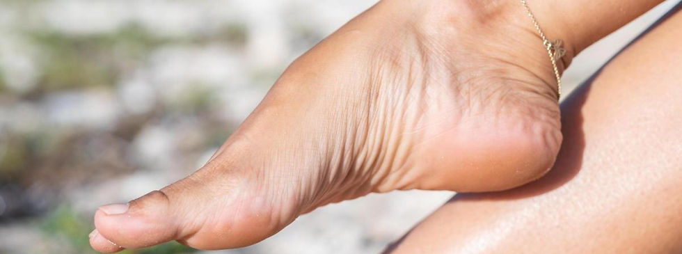 A sun-tanned right foot and ankle wearing a delicate metal chain anklet, resting horizontally against smooth, gray river stones.