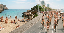 An ultra-wide widescreen split photograph showing a scenic, sandy beach cove where people are sunbathing and swimming clothes-free on the left, blending into a wide-angle view of hundreds of body-painted, clothes-free cyclists riding down a coastal city boulevard on the right.