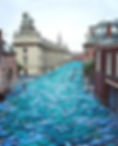 A high-angle photograph looking down a long city street flanked by grand historical stone and brick buildings, where thousands of unclad people painted in vibrant shades of blue and sea-green are lying side-by-side on the pavement to resemble a river of water.